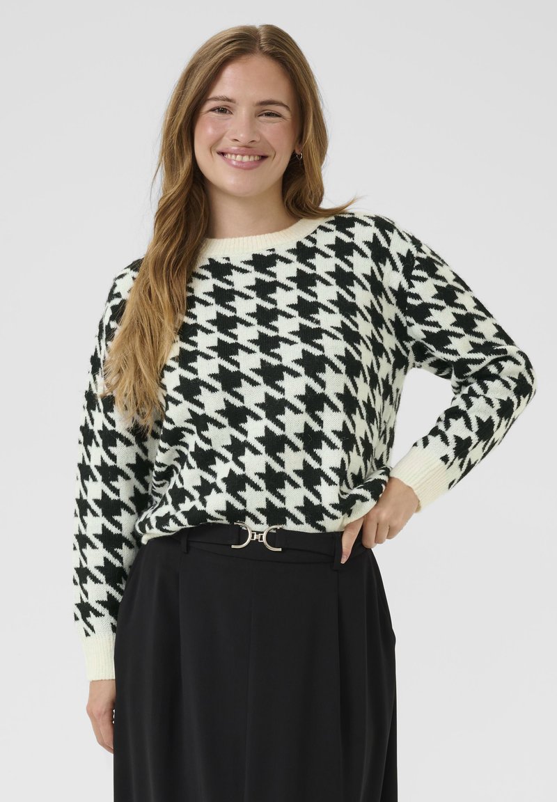 Black and white houndstooth-patterned knitted sweater with a round neckline and ribbed cuffs, worn over high-waisted black trousers with a belt.