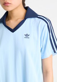 Svetlo modra polo majica s kratkimi rokavi, s temnim ovratnikom in tremi temnimi črtami na ramenih. Adidas logo v temno modri barvi na levem prsnem prostoru.