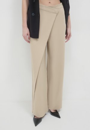 Beige wide-leg pants with overlapping front panel, worn with black sleeveless blazer and black pointed-toe shoes on a plain background.