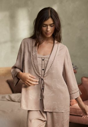 Frau trägt beige Pyjama-Set mit Blumenmuster und Bindevorne Jacke, steht in einem sanft beleuchteten Schlafzimmer mit neutralen Farben.