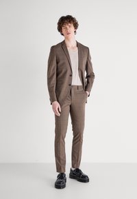 Costum maro cu carouri, cu o sacou croit și pantaloni slim-fit, asortat cu o bluză bej deschis și pantofi cu platformă negri.
