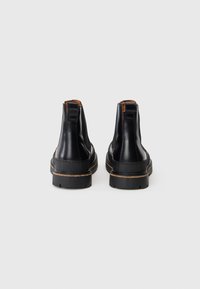 Une paire de bottes Chelsea en cuir noir avec des détails marron, vues de l'arrière sur un fond blanc uni.