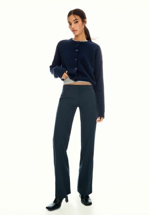 Femme portant un cardigan bleu marine sur un haut gris, un pantalon bleu marine à rayures et des bottes à talons noires, se tenant devant un fond blanc.