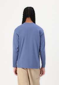 Chemise à manches longues en coton bleu doux, avec un col rond et une coupe décontractée. Coutures visibles et détail à l'ourlet arrondi.