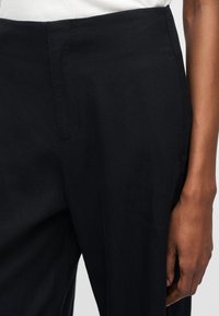 Κοντινό πλάνο μαύρου, ραμμένου παντελονιού, φορεμένου με λευκό τοπ, που δείχνει το πλάγιο ραφάκι, τη ζώνη μέσης και ένα χαλαρό χέρι που ακουμπάει δίπλα.