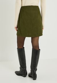 Mini-jupe en velours côtelé vert à côtes verticales, avec fermeture éclair au dos, associée à des bottes à talons hautes jusqu'au genou noires et des collants transparents.