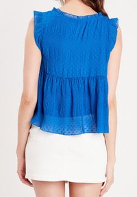 Haut bleu plissé avec manches courtes et accents à volants, en tissu léger et texturé. Associé à une mini-jupe blanche.