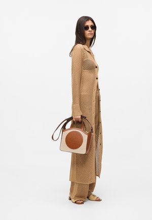 Žena u bež dugom kardiganu otvorenog pletenja i hlačama, drži bež i smeđu torbu, nosi tamne sunčane naočale i bež sandale.