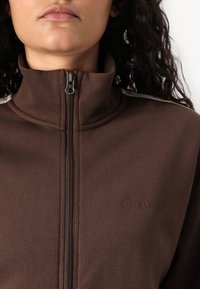 Brun zip-up jacka med hög krage, som har strukturerad tyg, en subtil logotyp och flerfärgade accentränder på axlarna.