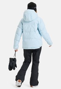 Lichtblauwe pufferjack met een capuchon, geborduurde ontwerp en synthetisch materiaal, gecombineerd met zwarte sneeuwbroeken en zwarte handschoenen, staande op sneeuw.