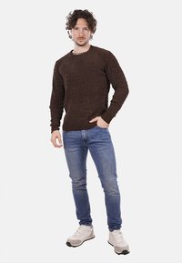 Maglione marrone a maglia texture con scollatura rotonda, abbinato a jeans slim-fit blu chiaro e sneaker bianche con dettagli neri.