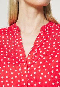 Czerwona bluzka z krótkim rękawem i kołnierzykiem, zdobiona białym wzorem w kropki. Tekstura wydaje się lekka i przewiewna.