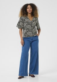 Blouse noire et crème à motif avec un col en V et des manches courtes, accompagnée de jeans en denim bleus à jambes larges et de chaussures violettes foncées.