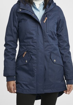 Kobieta ubrana w granatową, ocieplaną zimową kurtkę z wieloma kieszeniami na zamek i zatrzaski, nałożoną na jasnoniebieską koszulę i biały golf.