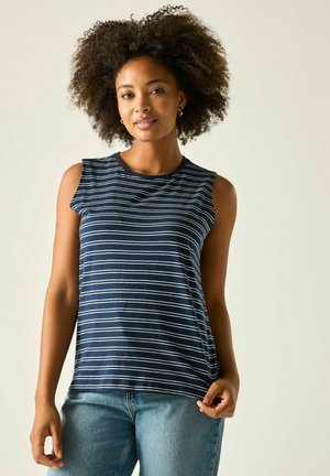 Mujer con pelo rizado que lleva una camiseta sin mangas a rayas azul marino y blanca y vaqueros azules sobre un fondo liso y claro.