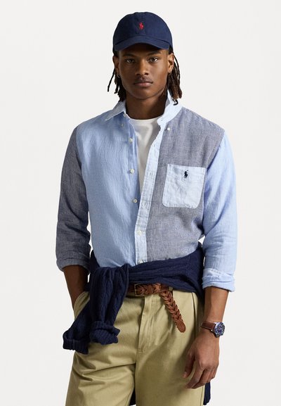 Lichtblauw en marineblauw gestreepte button-up shirt met een witte zak; marineblauwe pet; bruine gevlochten riem; kaki broek en een donkere trui geknoopt om de taille.