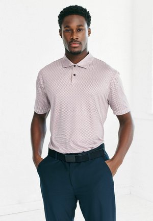 Kurzärmliges Polohemd in Hellrosa mit geometrischem Muster, kombiniert mit marineblauen Hosen und einem schwarzen Gürtel, vor einem weißen Hintergrund.