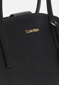 Sort håndtaske med tekstur og et reptilskindsmønster, guld "Calvin Klein"-logo og elegante håndtag for nem bæring.