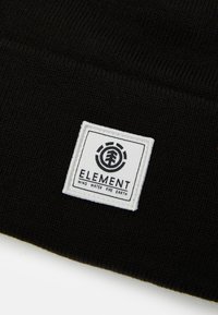 Tela de punto negra con un parche cuadrado blanco que muestra un logotipo de árbol circular y las palabras "ELEMENTO VIENTO AGUA FUEGO TIERRA."