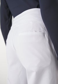 Pantalon blanc avec une texture lisse et un design de poche arrière. Présente un accent logo subtil près de la taille. Associé à un haut marine.