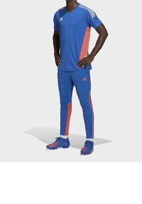 Modré sportovní tričko s růžovým bočním panelem a třemi bílými pruhy na ramenou, v kombinaci s modrými kalhotami s růžovými pruhy a logem.