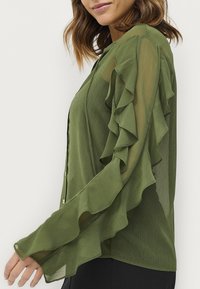 Oliven grøn bluse med lange, gennemsigtige, volangerede ærmer, knaplukning og let tekstureret stof. Moderne pasform med et flydende design.