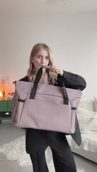 Růžová látková taška na rameno s černými detaily, horními rukojeťmi a bočními kapsami, zobrazena v rukou osoby na neutrálním pozadí.