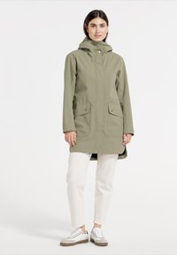 Olivgrön vattentät jacka med huva, två framfickor och tryckknappar, matchad med vita byxor och grå sneakers.