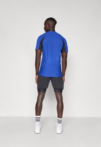 Camisa deportiva azul con diseño texturizado y detalles en negro, combinada con pantalones cortos negros. El modelo lleva zapatillas blancas y calcetas a rayas.