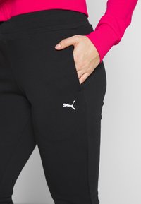 Personne portant un pantalon noir Puma avec la main dans la poche et un logo Puma blanc visible, associé à un haut à manches longues rose vif.
