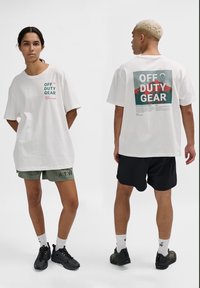 Vit oversized t-shirt med "OFF DUTY GEAR" tryckt i turkos och röd text. Stylar med gröna och svarta shorts. Texturerat bomullsmaterial.