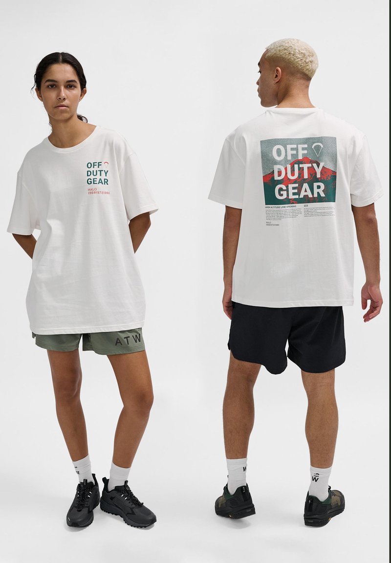 Vit oversized t-shirt med "OFF DUTY GEAR" tryckt i turkos och röd text. Stylar med gröna och svarta shorts. Texturerat bomullsmaterial.