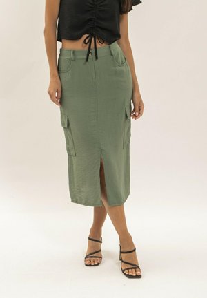 Femme portant un crop top noir froncé, une jupe midi vert olive fendue à l'avant avec poches latérales, et des sandales à talons noires à lanières.
