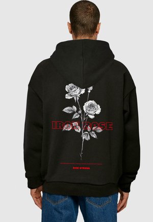Sort hættejakke med et blomstermotiv af hvide roser og teksten "IRON ROSE" i rødt. Den har en afslappet pasform og en kængurulomme.