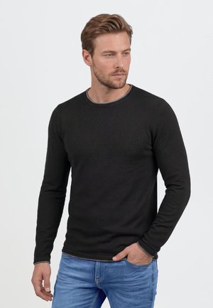Homme aux cheveux châtain clair et barbe, portant une chemise noire à manches longues et un jean bleu, debout devant un fond blanc uni.
