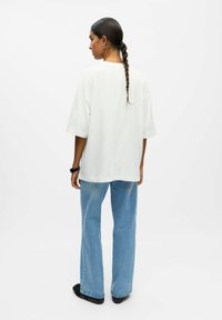 Bijela oversized majica od pamuka, uparena s svijetloplavim farmerkama s mašnom. Model ima pletenu frizuru i nosi crne cipele.