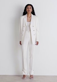 Weiße, taillierte Blazerjacke mit doppeltknöpfiger goldener Knopfleiste, kombiniert mit passenden, figurschmeichelnden Hosen und einem hellen Oberteil. Getragen mit offenen Schuhen.