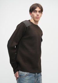 Barna pulóver, texturált kötésű, fekete bőr vállrészletekkel, kerek nyakkal és egy kis mellzsebbel. Kék farmernadrággal viselve.