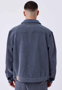 Giacca a quadri blu con tessuto strutturato, caratterizzata da un colletto e maniche con bottoni. Un'etichetta è attaccata vicino all'orlo.