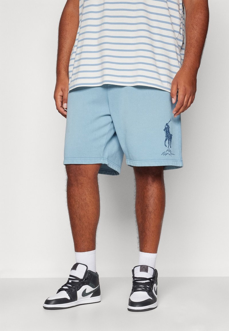 Světle modré bavlněné šortky s elastickým pasem, bočními kapsami a grafickým logem na levé noze. Vzhledem k černobílým teniskám.