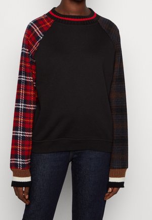 Personne portant un sweat-shirt noir avec des manches à carreaux dépareillées : rouges et blanches à gauche, tons foncés à droite, et des poignets côtelés multicolores.