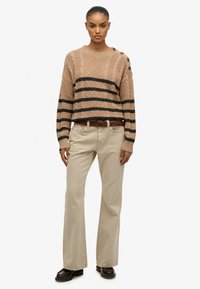 Beige kabelbrei sweater met zwarte strepen, met knoopaccenten op de schouder. Gecombineerd met lichtgekleurde flared broeken en een bruine riem.