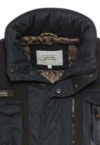 Marineblå pufferjakke med høj krave, der har et mønstret indre, frontlynlås, trykknapper og sidelommer for ekstra funktionalitet.