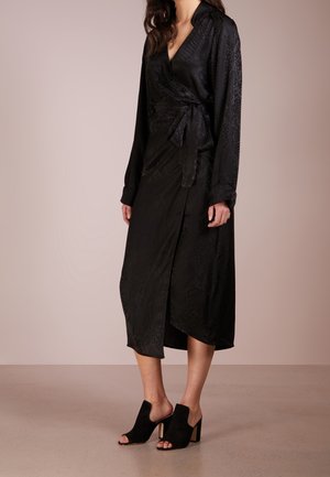 Kvinde iført sort omslagskjole med lange ærmer og diskret mønster, med bælte i taljen, og sorte åbne hælede mules, stående på neutral baggrund.