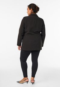 Juodas švarkas su baltomis taškeliais, derinamas su juodomis leginsais, turinčiais panašų raštą, ir sidabro akcentą turinčiais smailiais batais. Lygus audinio tekstūra.