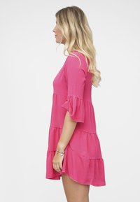 Lyhythihainen vaaleanpunainen mekko, jossa on kerroksittainen muotoilu ja röyhelökoristelu, valmistettu kevyestä, laskeutuvasta kankaasta. Sivukuva esittää mekon pituutta ja siluettia.