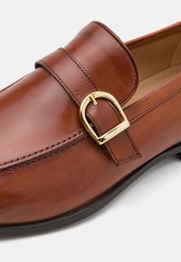 Braune Lederloafers mit einem Riemen und goldfarbenem Schnallen-Detail. Sie verfügen über eine glatte Textur und eine abgerundete Zehenform und ruhen auf einer schwarzen Gummisohle.