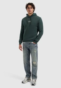 Donkergroen hoodie met verstelbare trekkoord, voorzien van een logoprint aan de voorkant. Gecombineerd met lichtblauwe distressed jeans en zilveren sneakers.