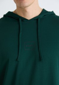 Hanorac verde din material moale, cu un buzunar tip kangaroo, glugă cu șnur și un logo pătrat "HUGO" în negru pe piept.