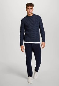 Marineblauw kabelgebreide trui met een ronde halslijn, gecombineerd met donkere broek en witte sneakers, waardoor een gelaagde look met textuur ontstaat.